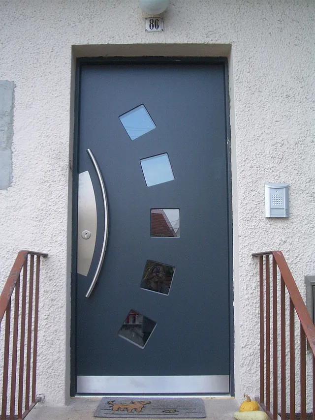 Dunkelgraue Haustür mit fünf schräg angeordneten quadratischen Fenstern und langem, gebogenem Griff aus Metall.