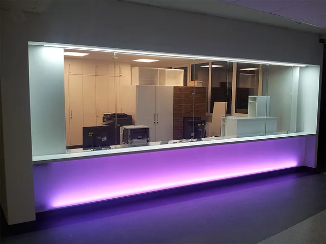 Blick durch eine große Glasscheibe auf einen Raum mit weißen Schränken und Computermonitoren, davor eine Theke mit lila Beleuchtung.