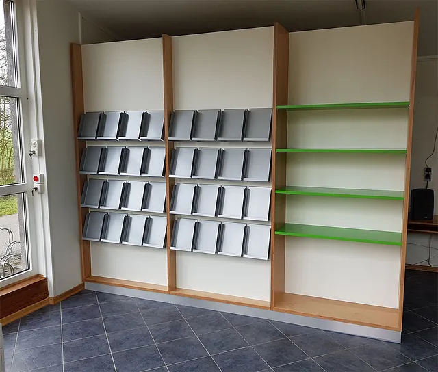 Leeres Holzregal mit grünen Regalböden auf der rechten Seite und grauen, schrägen Zeitschriftenständern auf der linken Seite. Der Raum hat einen Fliesenboden und ein großes Fenster.