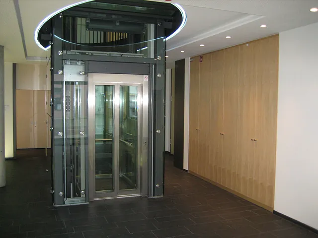 Glasaufzug mit Metallrahmen in einem Flur mit dunklem Boden und hellen Wänden.