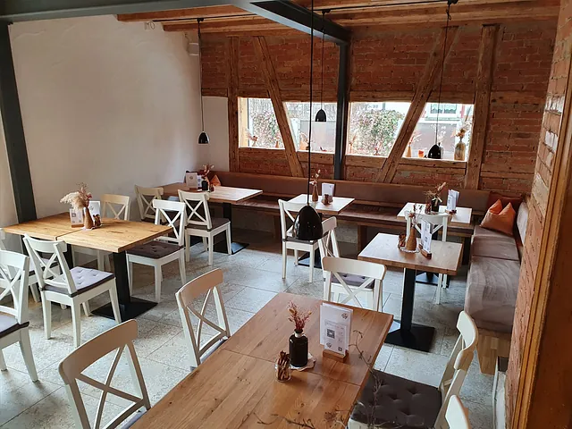 Gastronomieraum mit Holztischen, weißen Stühlen und Sitzbank, dekoriert mit kleinen Vasen und Menükarten, Fenster mit Blick nach draußen.