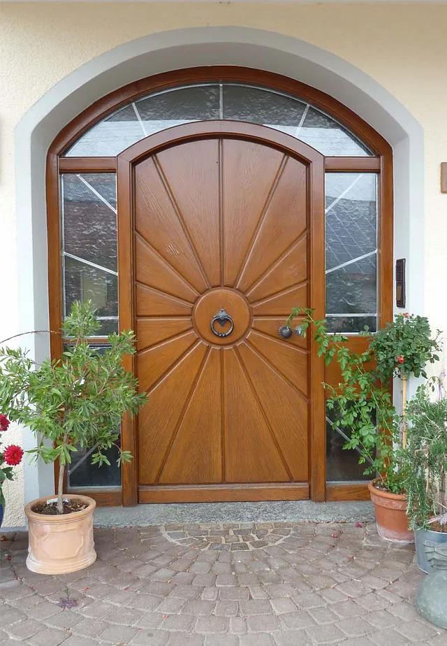 Holztür mit rundem Türklopfer und strahlenförmiger Musterung, umgeben von Glasfenstern und Pflanzen in Töpfen.