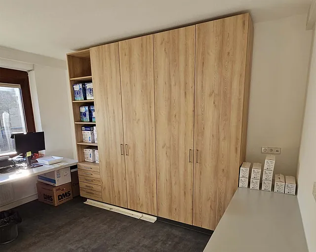 Schrank mit Holzoptik und mehreren Türen in einem Raum mit Fenster, Schreibtisch und Kartons.