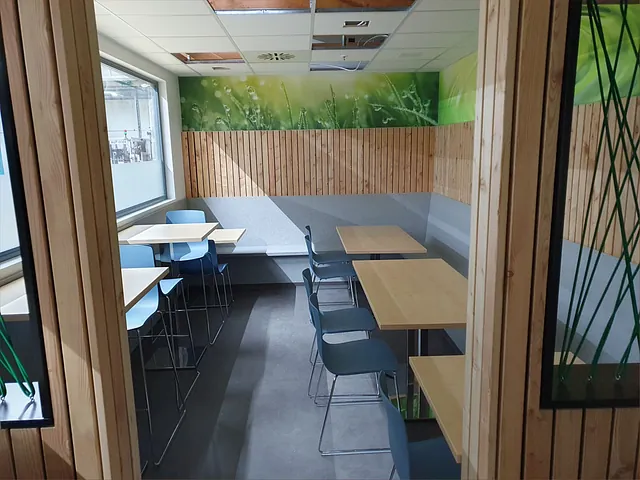 Kantinenraum mit Holzwandverkleidung, grüner Wandgestaltung, Tischen und blauen Stühlen, Blick durch Glastür.