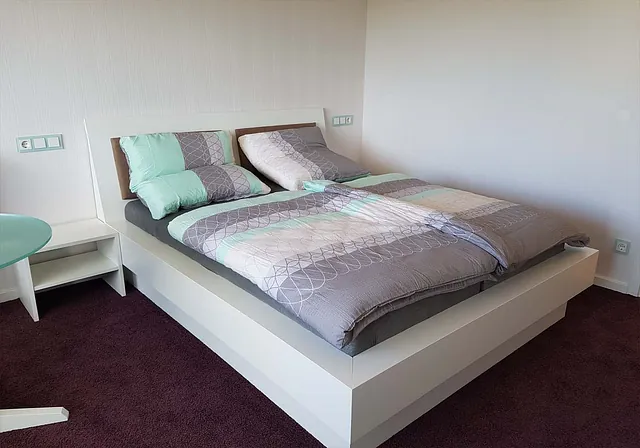 Modernes Doppelbett mit weißem Rahmen, grauer Matratze und Bettwäsche in Grau- und Minttönen mit geometrischem Muster, daneben weißer Nachttisch und runder mintfarbener Tisch auf dunkelrotem Teppich.