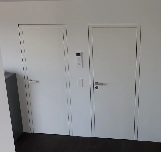 Zwei weiße Türen mit silbernen Griffen an einer weißen Wand, dazwischen ein Thermostat und ein Lichtschalter.