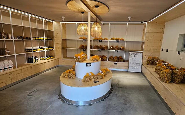 Moderner Laden mit Holzregalen, in denen Kartoffelsäcke und verschiedene Produkte ausgestellt sind. Mit zentraler Insel mit zwei geflochtenen Lampen.