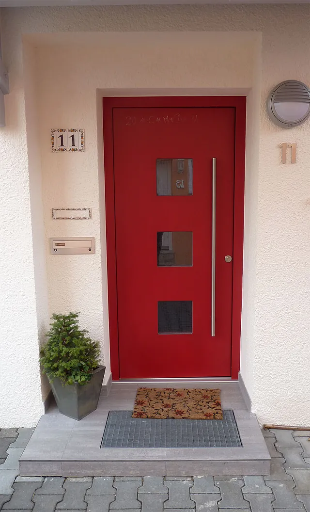 Rote Haustür mit drei quadratischen Fenstern und langem Griff, daneben Hausnummer 11, Fußmatte und Pflanze in grauem Topf