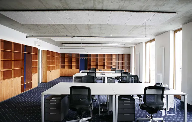 Großer Büroraum mit mehreren weißen Schreibtischen, schwarzen Bürostühlen und leeren Holzregalen an den Wänden.