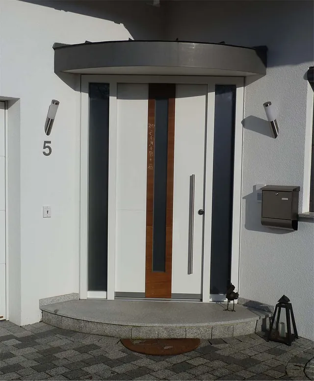 Eingangstür mit vertikalem Holzstreifen und zwei schmalen Fenstern, Hausnummer 5, Briefkasten und Laterne auf der rechten Seite