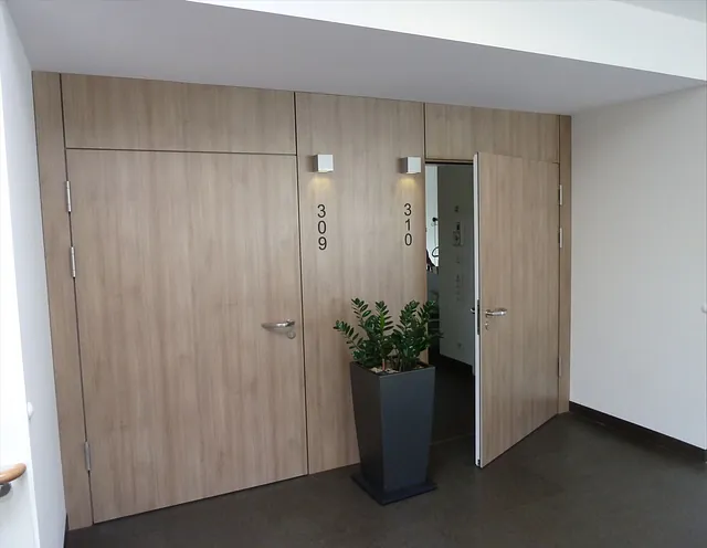 Zwei Holztüren mit den Nummern 309 und 310 in einem Flur, wobei die Tür zu Zimmer 310 teilweise geöffnet ist. Zwischen den Türen steht eine Topfpflanze.