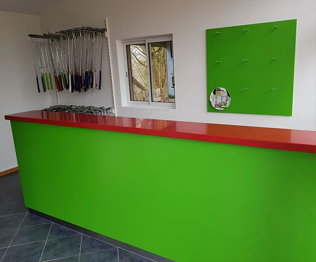 Grüner Tresen mit roter Arbeitsfläche in einem Raum, an der Wand ein grünes Brett mit Haken und ein Fenster, daneben hängen Golfschläger.