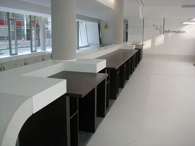 Moderner Informationsschalter in elegantem Design aus weißem und dunklem Holz, gelegen in einem geräumigen, hellen Raum mit großen Fenstern. Minimalistisch und einladend.