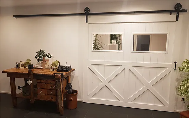 Weißes Schiebetor mit zwei rechteckigen Fenstern neben einem rustikalen Holztisch mit Schreibmaschine und Pflanzen.