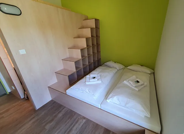 Zwei Einzelbetten mit weißen Bettdecken und Handtüchern, daneben ein Treppenregal aus hellem Holz, grüne Wand im Hintergrund.