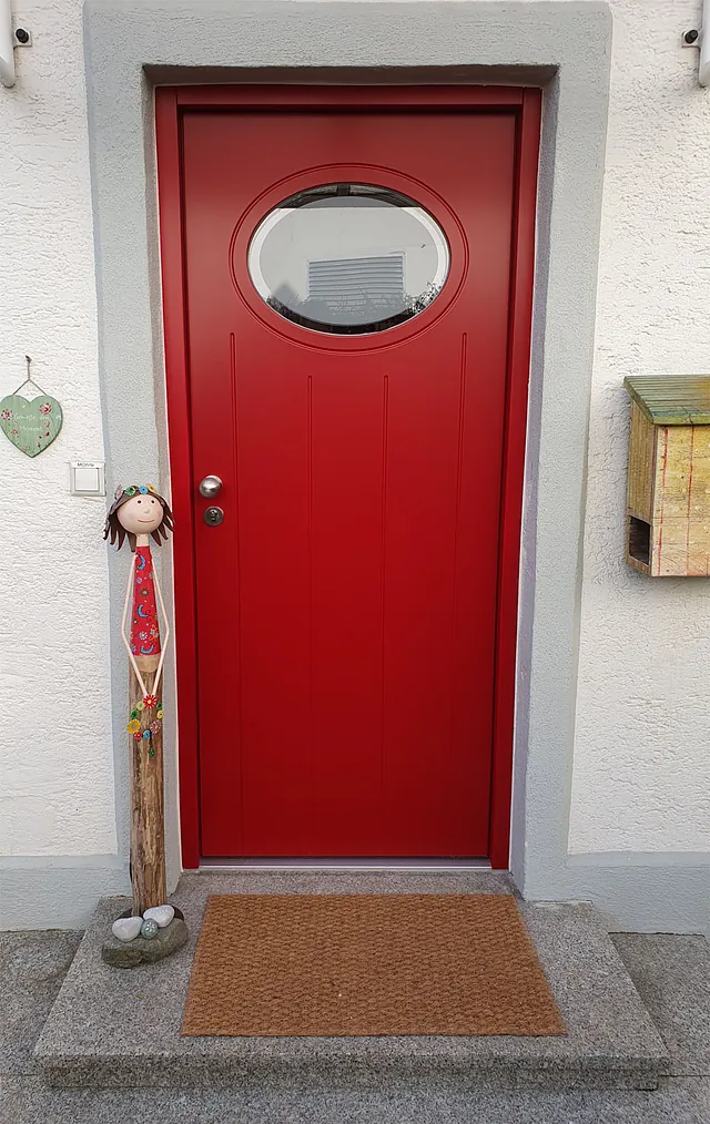 Rote Haustür mit ovalem Fenster, daneben eine Holzfigur mit rotem Kleid und braunen Haaren, Fußmatte vor der Tür.